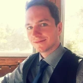 Smiling man with blue eyes, wearing a vest and tie, bright window behind him.